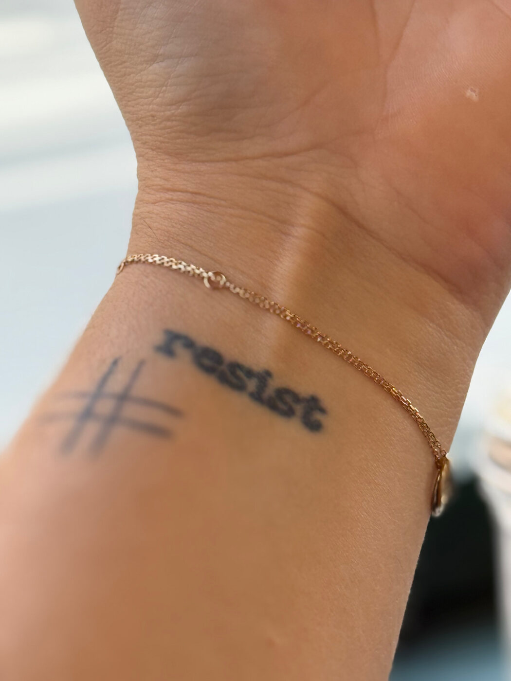 A close view of a wrist bearing a handwritten-style Resist tattoo and a delicate gold bracelet, symbolizing equality and civic action