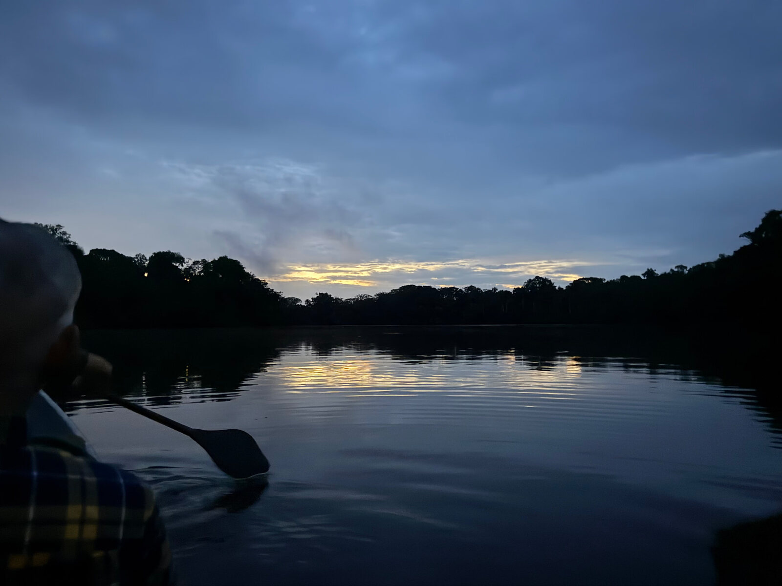 A person paddles a canoe across calm water at dusk in Ecuador, surrounded by a quiet blue landscape and the last light on the horizon